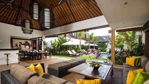 A modern living room with a large sofa, a coffee table, and a dining area with chairs and a table. There is a view of a pool and outdoor seating with umbrellas. - Bali Villa Near the Beach, 2061 (Seminyak)