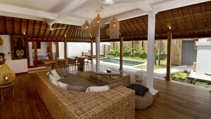 A spacious living area with a large wicker sofa, a dining table, and chairs. There is a ceiling fan, hanging lights, and a view of a pool and garden outside. - Villa for Rent in Bali 2039 (Seminyak)