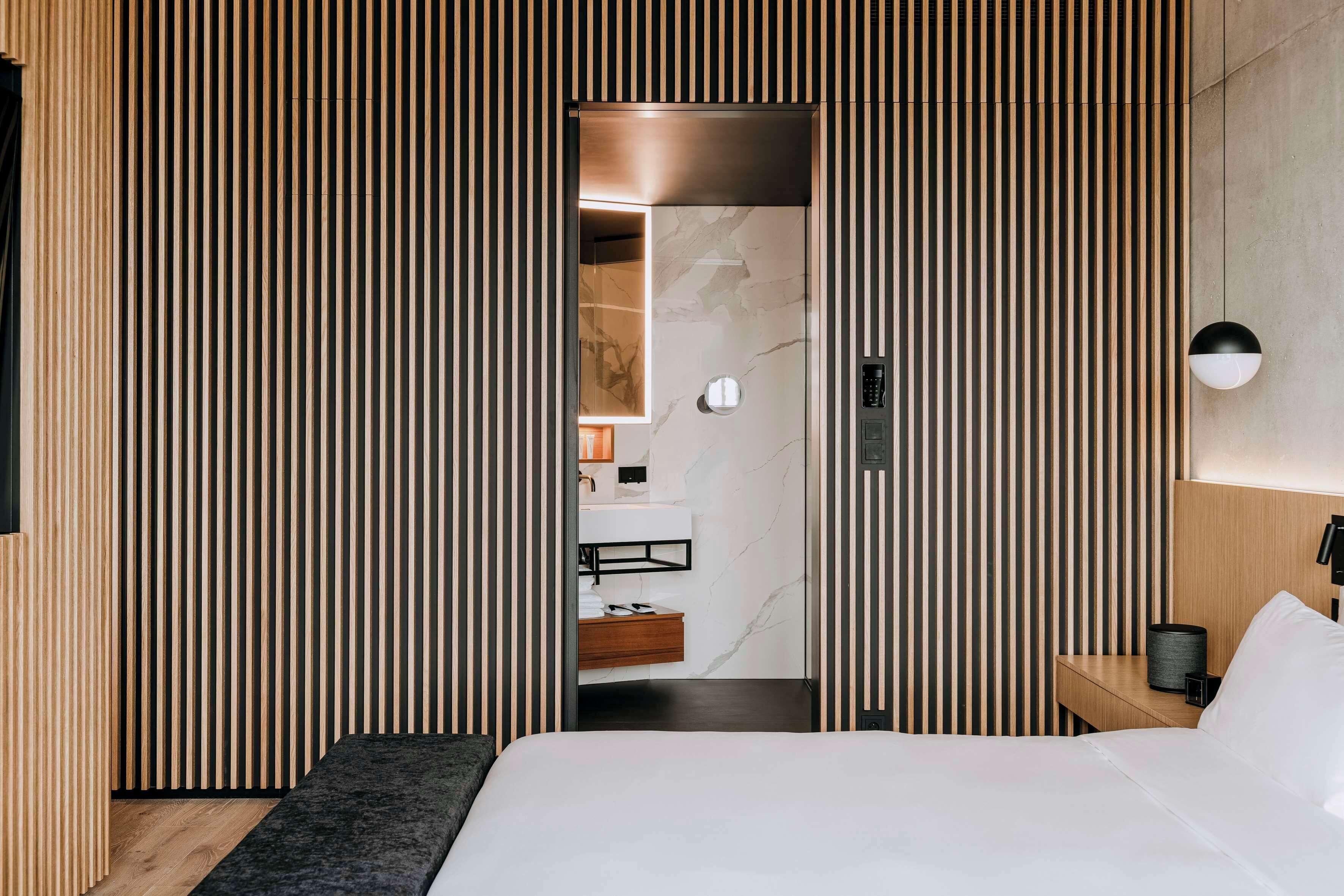 Una habitación de hotel moderna con cama, mesita de noche, lámpara de pared y diseño de paneles de madera.