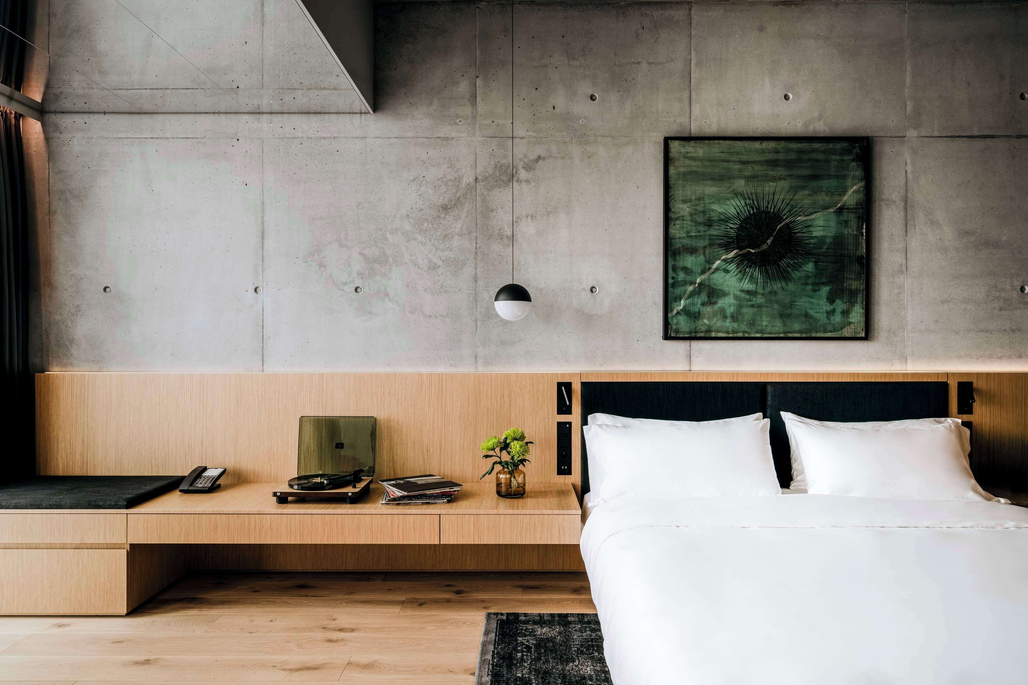 A modern bedroom with a wooden bedside table, a bed with white linens, a green abstract painting, and a small plant.