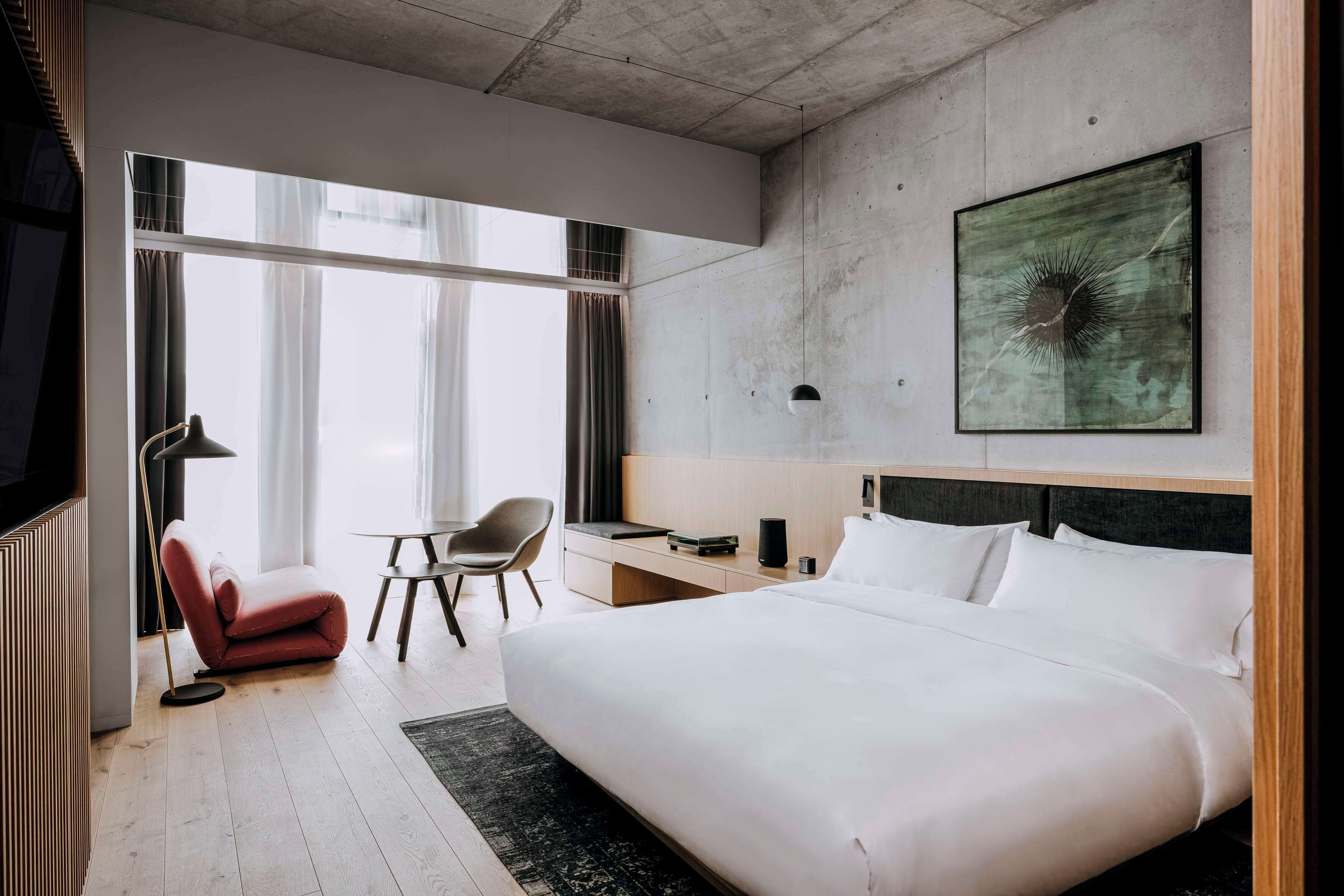A modern hotel room with a large bed, a desk, a chair, and a large window.