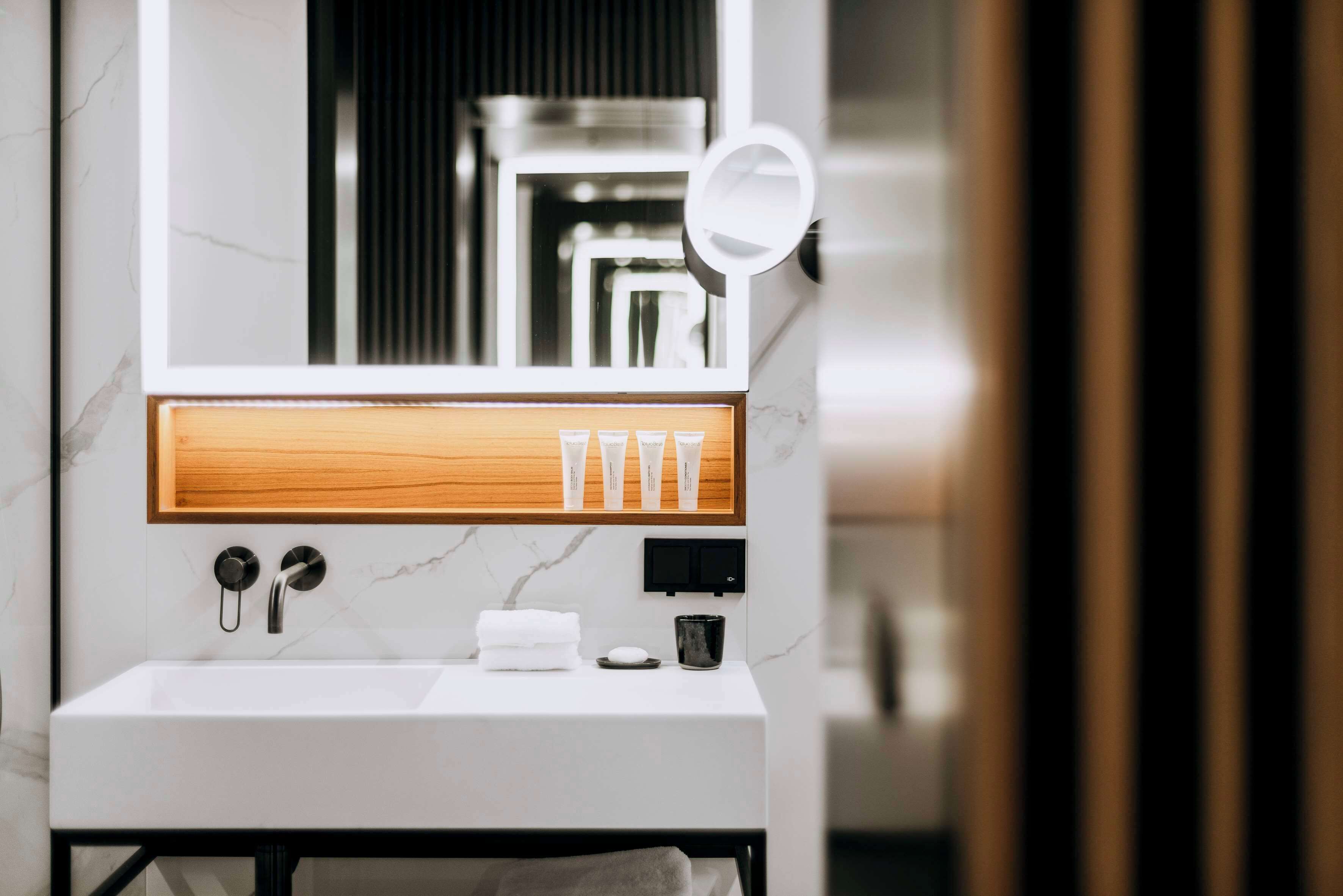 Une salle de bain moderne avec un lavabo blanc, un meuble-lavabo en bois, un miroir et une étagère contenant des articles de toilette.