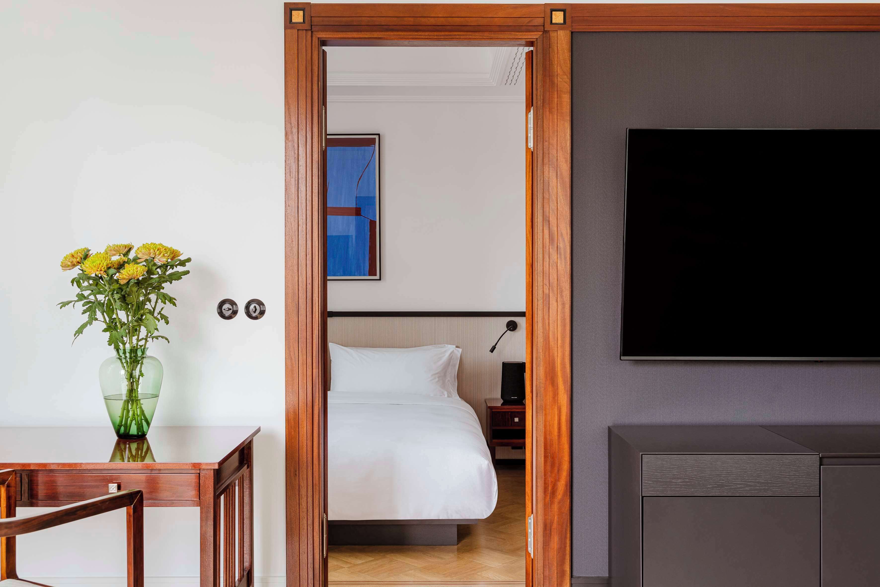 A hotel room with a wooden door, a bed with white bedding, a TV mounted on the wall, and a small table with a vase of flowers.
