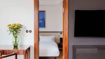 A hotel room with a wooden door, a bed with white bedding, a TV mounted on the wall, and a small table with a vase of flowers.