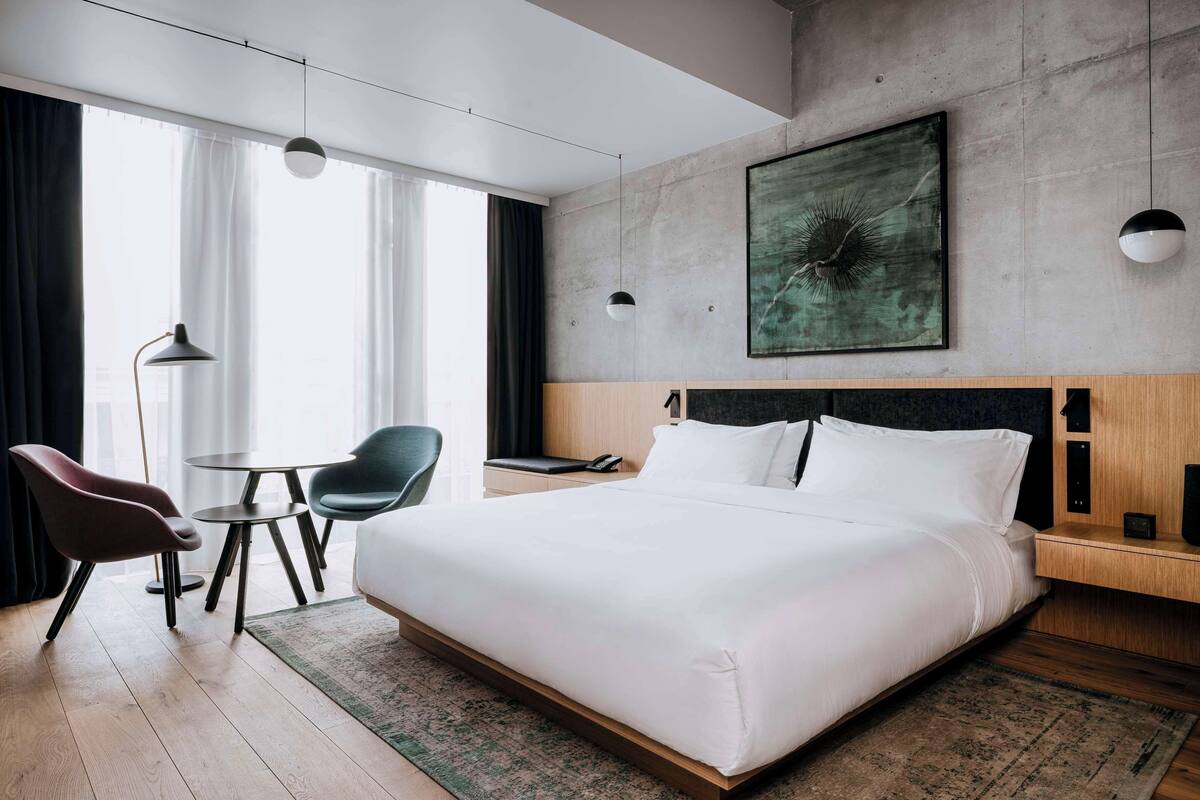 A modern hotel room with a large bed, a desk, two chairs, a round table, and a large window with curtains.