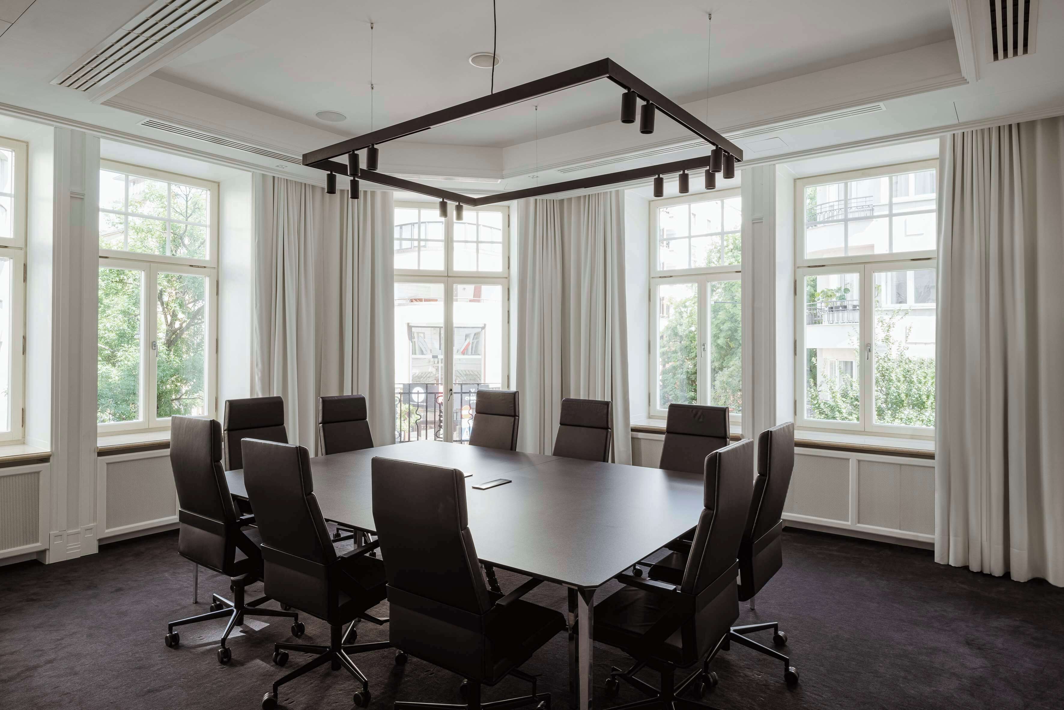A conference room with a large table and eight chairs, large windows with curtains, and a modern ceiling design.