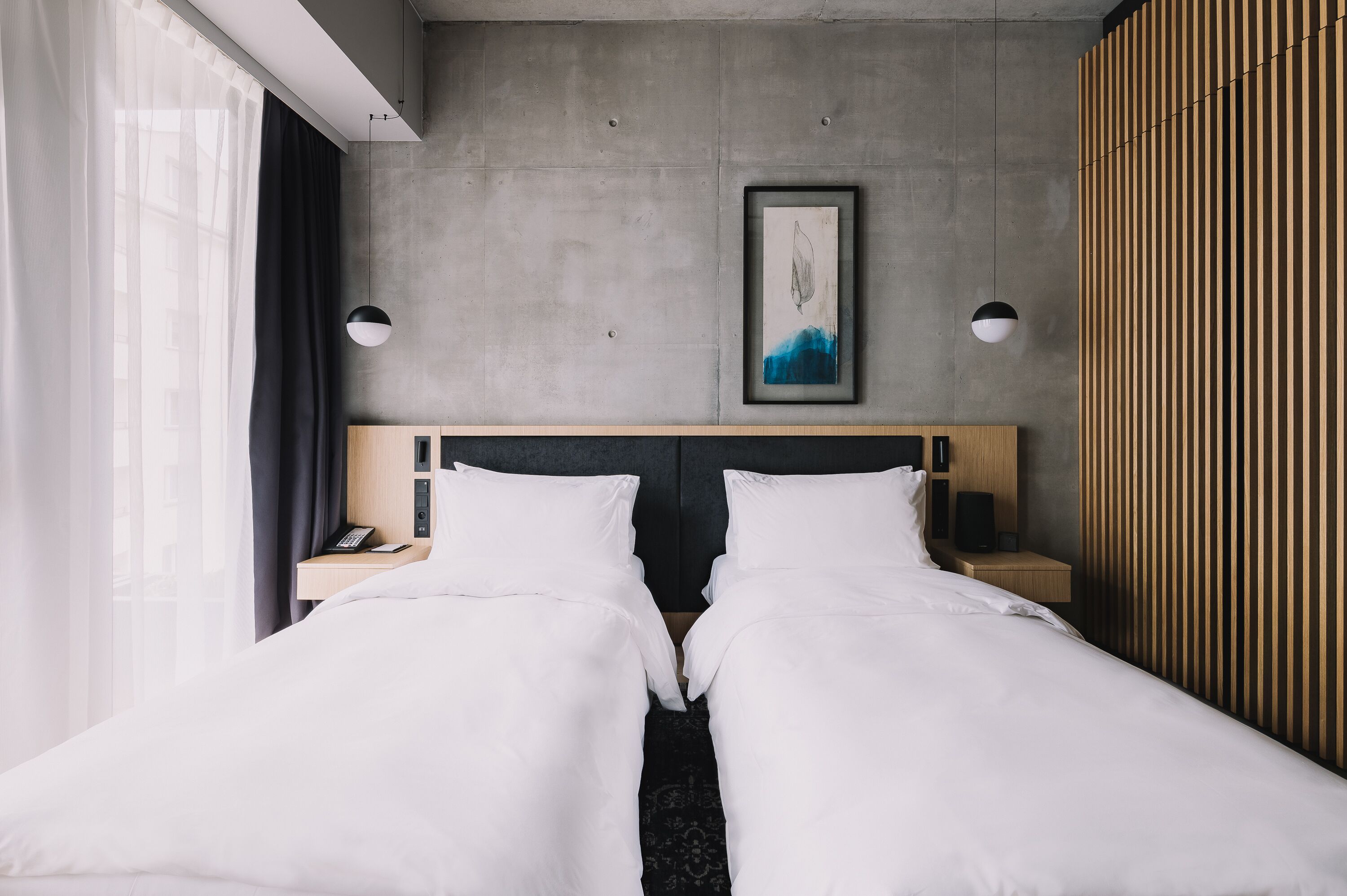 A hotel room with two beds, a large window with sheer curtains, a painting on the wall, and a wooden panel on the right.