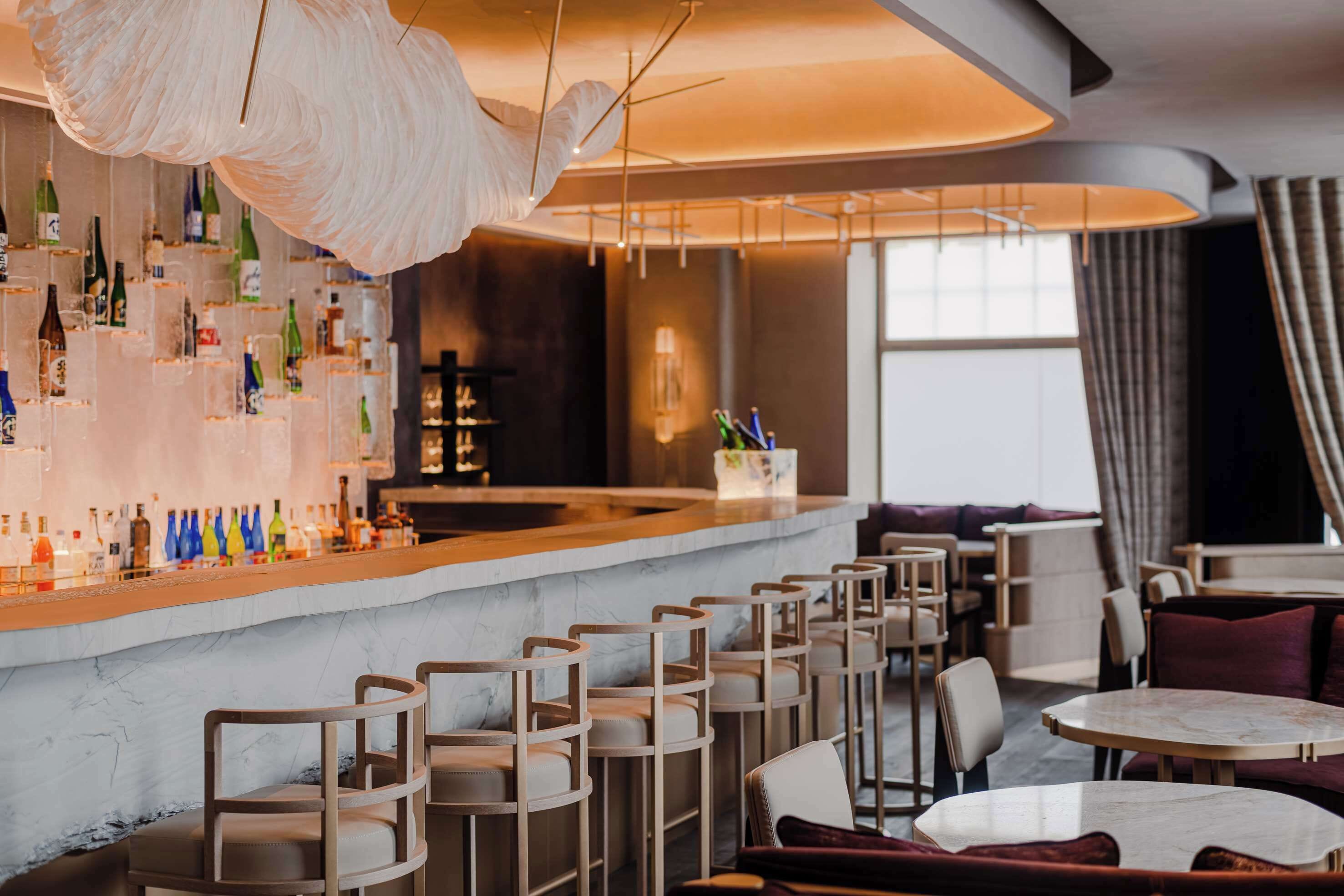 A bar area with a marble countertop, multiple seating options including bar stools and tables, and a variety of bottles on the shelves.