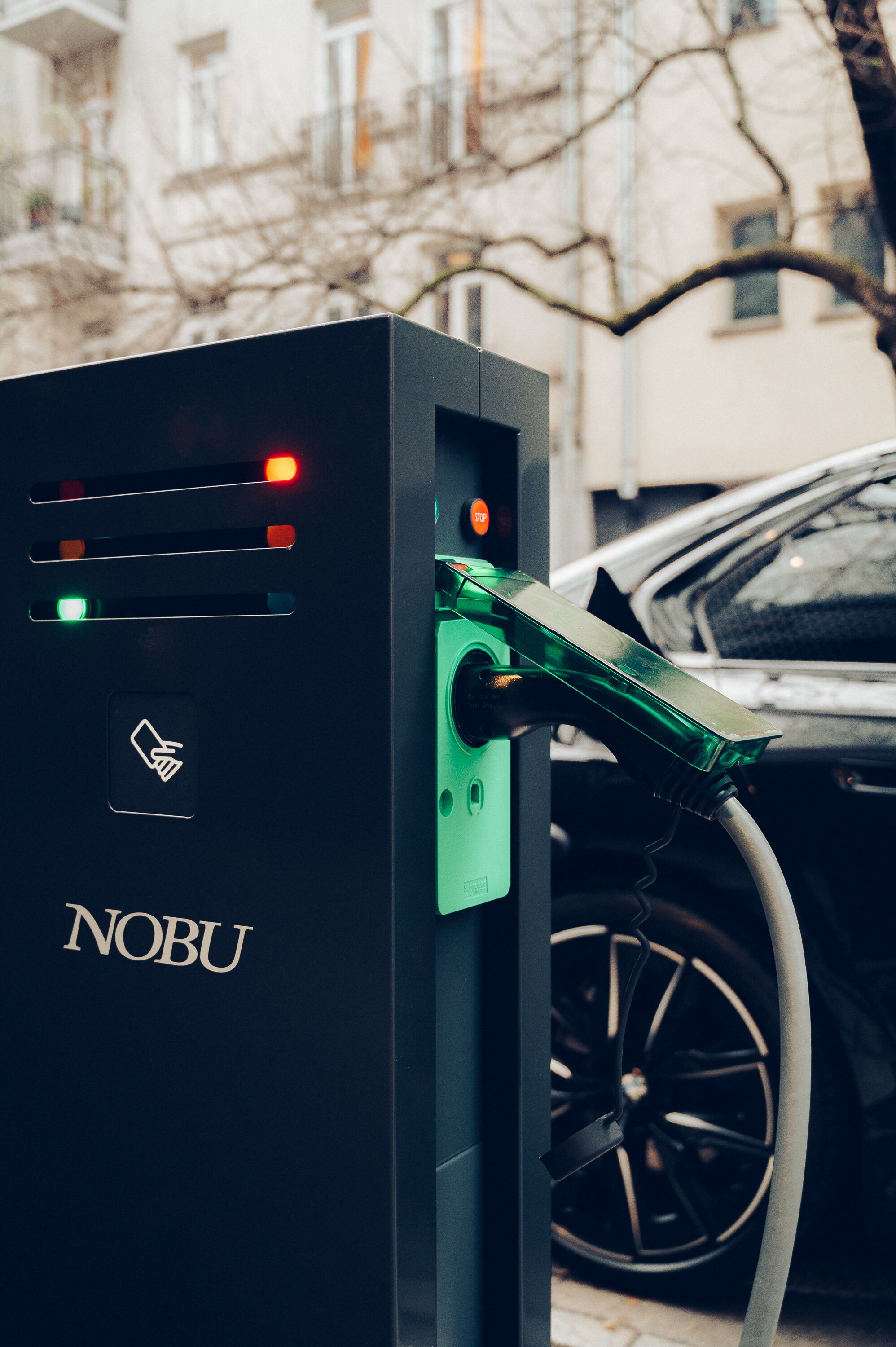 A black electric vehicle charging station with a green charging connector and a car parked nearby.