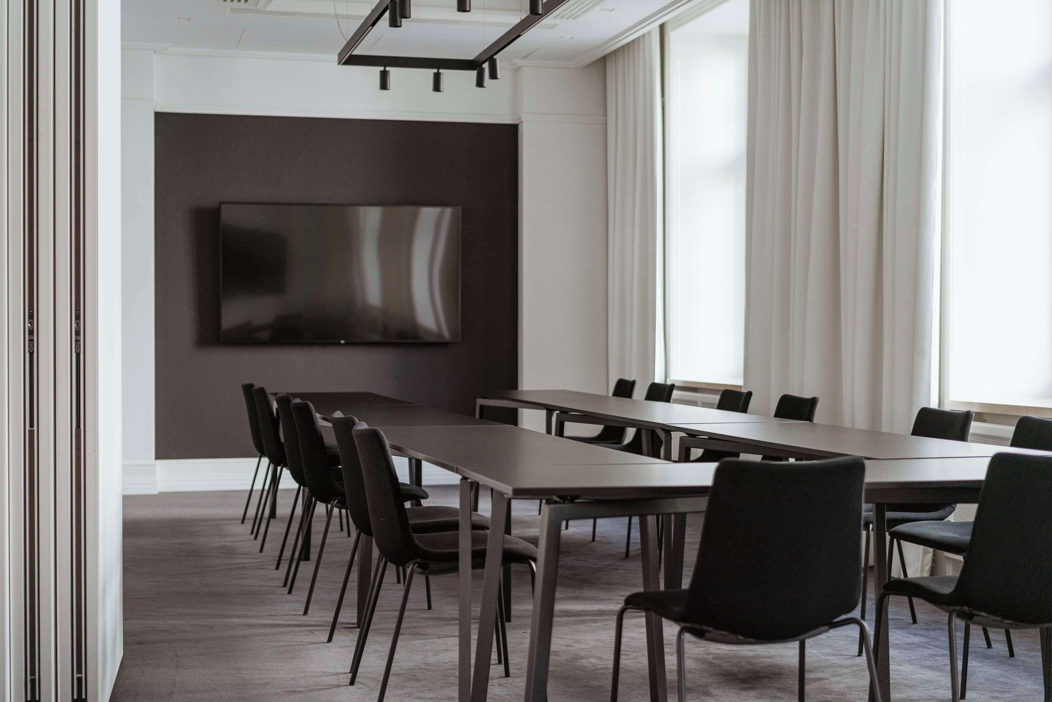 Una sala de conferencias con una mesa larga, sillas negras, un televisor de pantalla plana en la pared y ventanas amplias con cortinas.