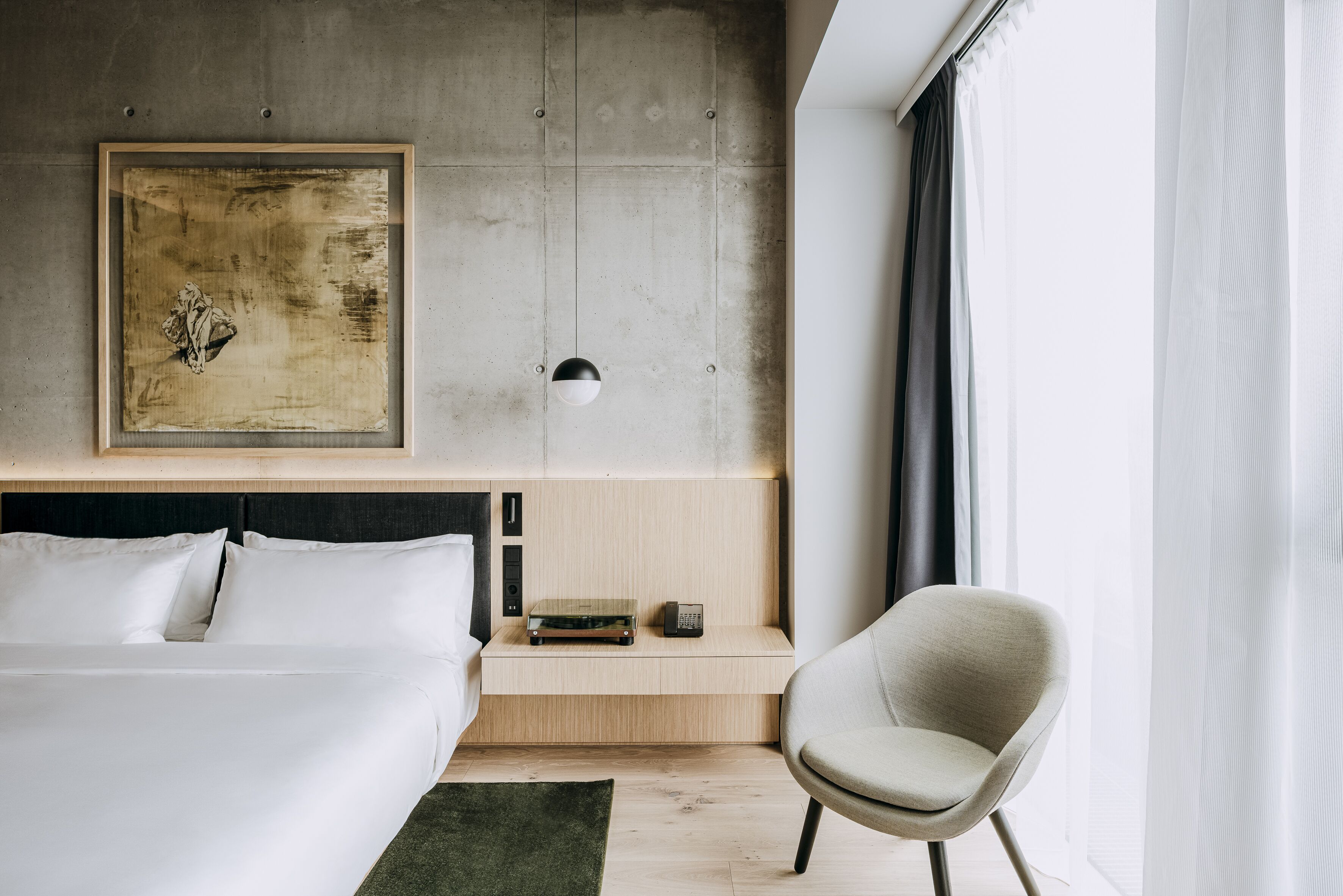 A modern hotel room with a large bed, a bedside table with a lamp, a chair, and a large abstract painting on the wall.