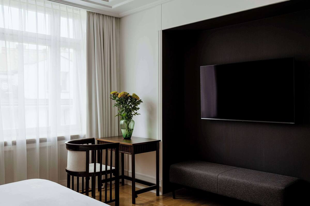 A hotel room with a bed, a desk with a chair, a vase with flowers, and a large flat-screen TV mounted on the wall.