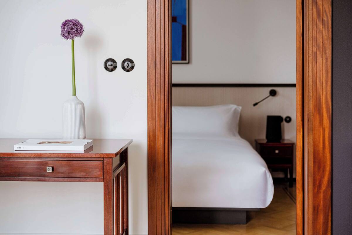 A hotel room with a wooden desk, a white vase with a purple flower, a white bed, a nightstand, and a wall-mounted lamp.