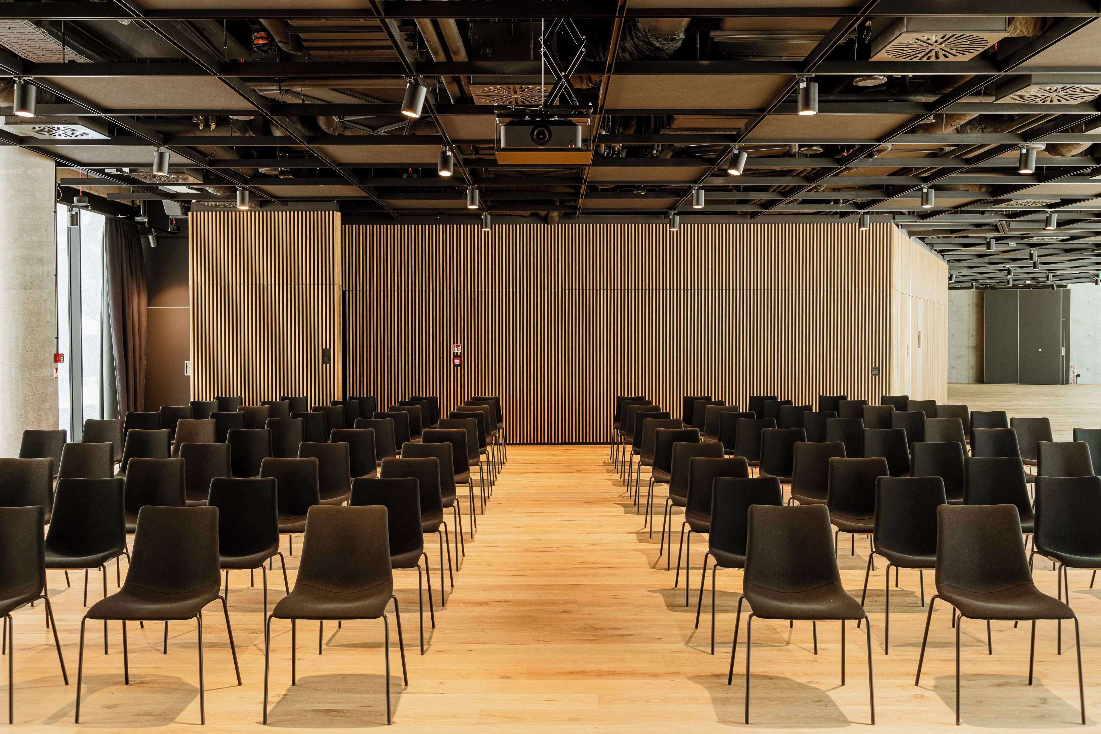 A conference room with rows of black chairs facing a wooden slat wall and a projector screen at the front.