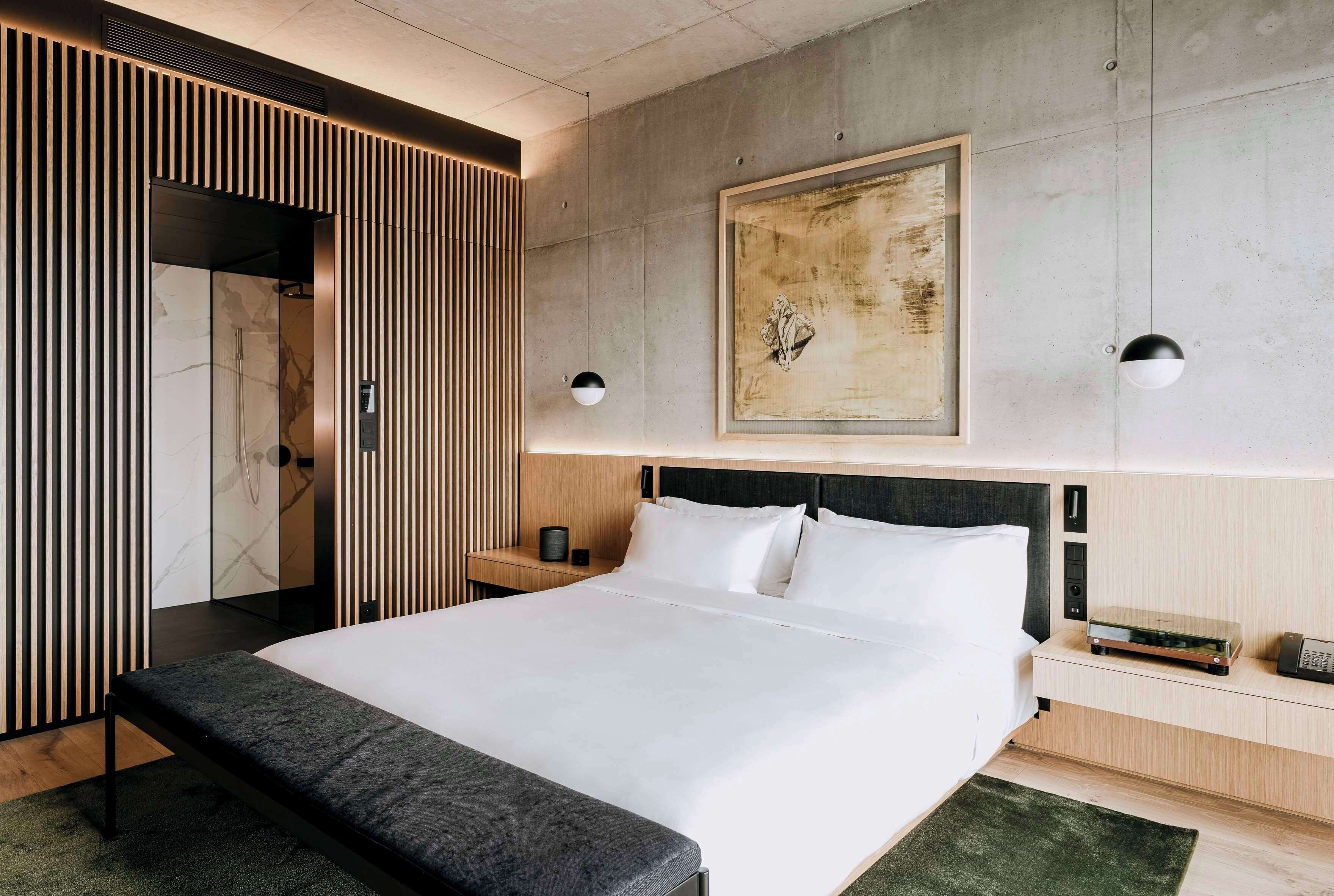 A modern hotel room with a large bed, a bedside table with a lamp, a wall-mounted artwork, and a wooden panel design on the left wall.