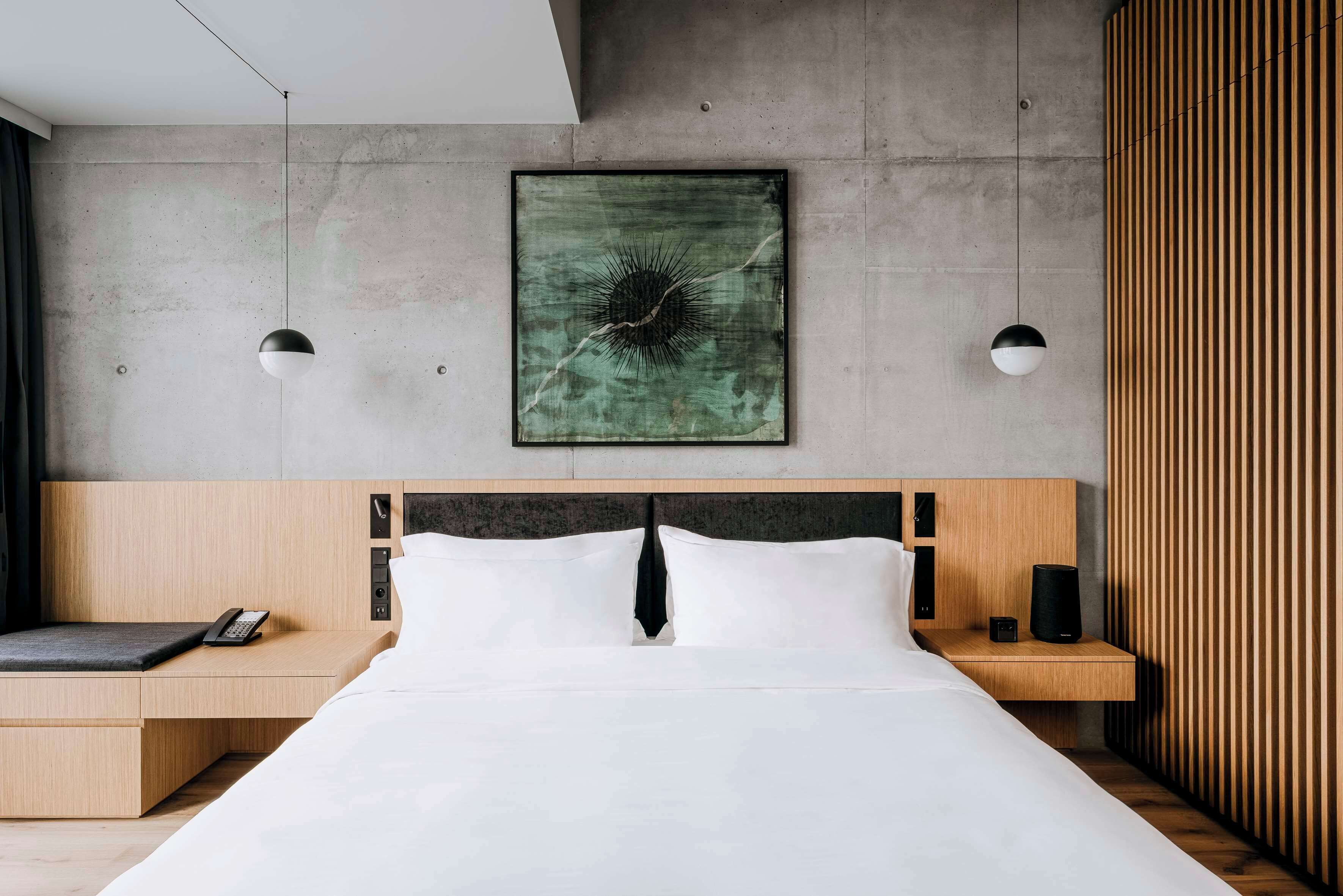 A modern hotel room with a large bed, a wooden nightstand with a phone, a wall-mounted artwork, and a wooden panel wall.