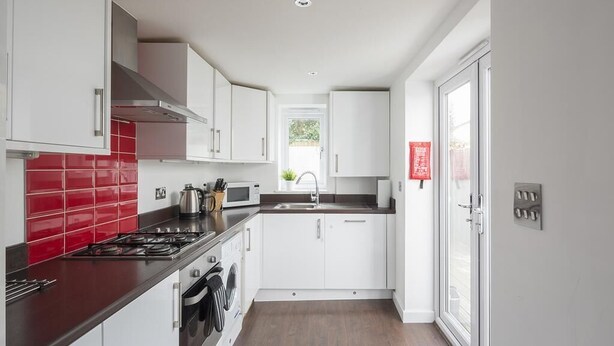 A modern kitchen with white cabinets, a red backsplash, stainless steel appliances, and a window providing natural light. - Sunny Terrace (Maidstone)