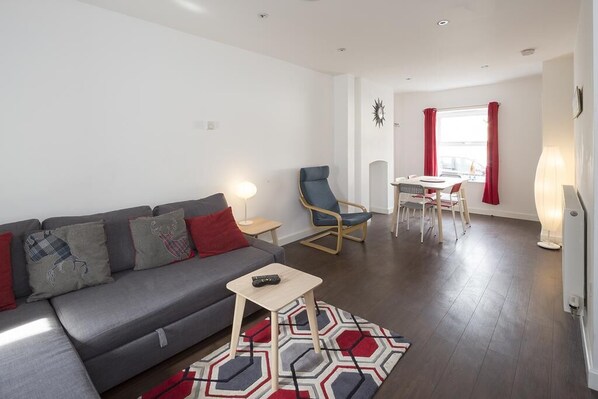 A modern living room with a grey sofa, a small wooden table, and a blue armchair. A dining area with a table and chairs is visible in the background.