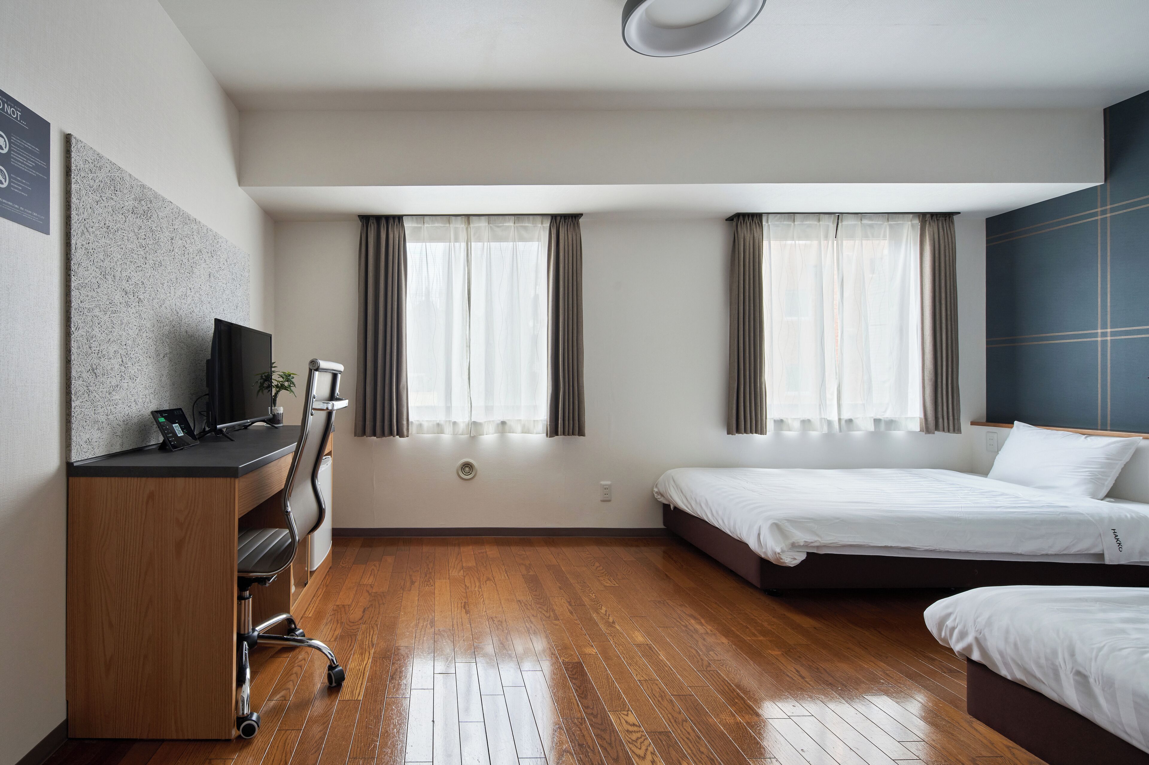 A hotel room with a wooden desk, two beds, and a large window with curtains.