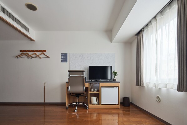 A modern hotel room with a wooden floor, a desk with a TV, a chair, a small cabinet, and a window with curtains.