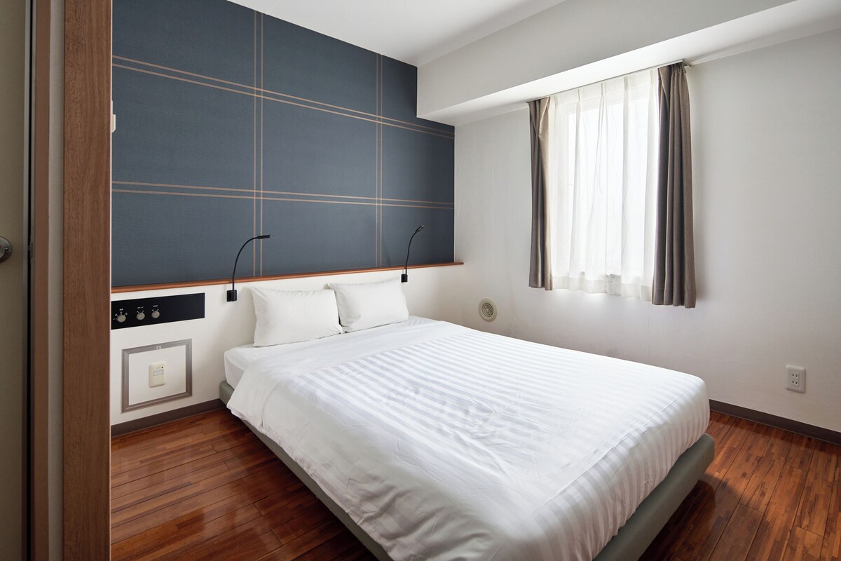 A modern hotel room with a large bed, wooden flooring, a built-in TV, and a window with curtains.