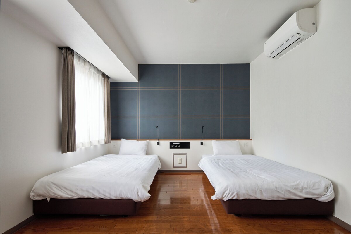 A hotel room with two single beds, a large window with curtains, a wall-mounted air conditioner, and a control panel on the wall.