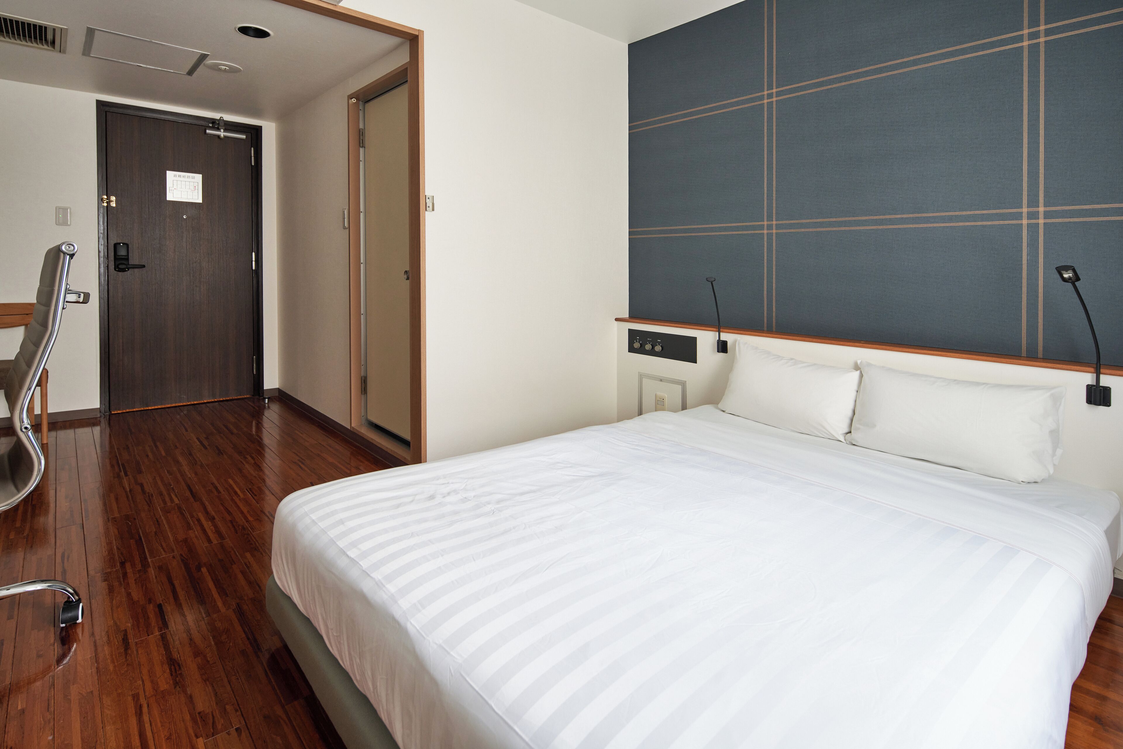 A hotel room with a large bed, a wooden floor, a dark-colored door, and a wall with a grid pattern.
