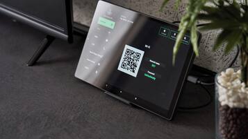 Een tablet toont een QR-code en Wi-Fi instellingen, met een plant en een kabel op de achtergrond.