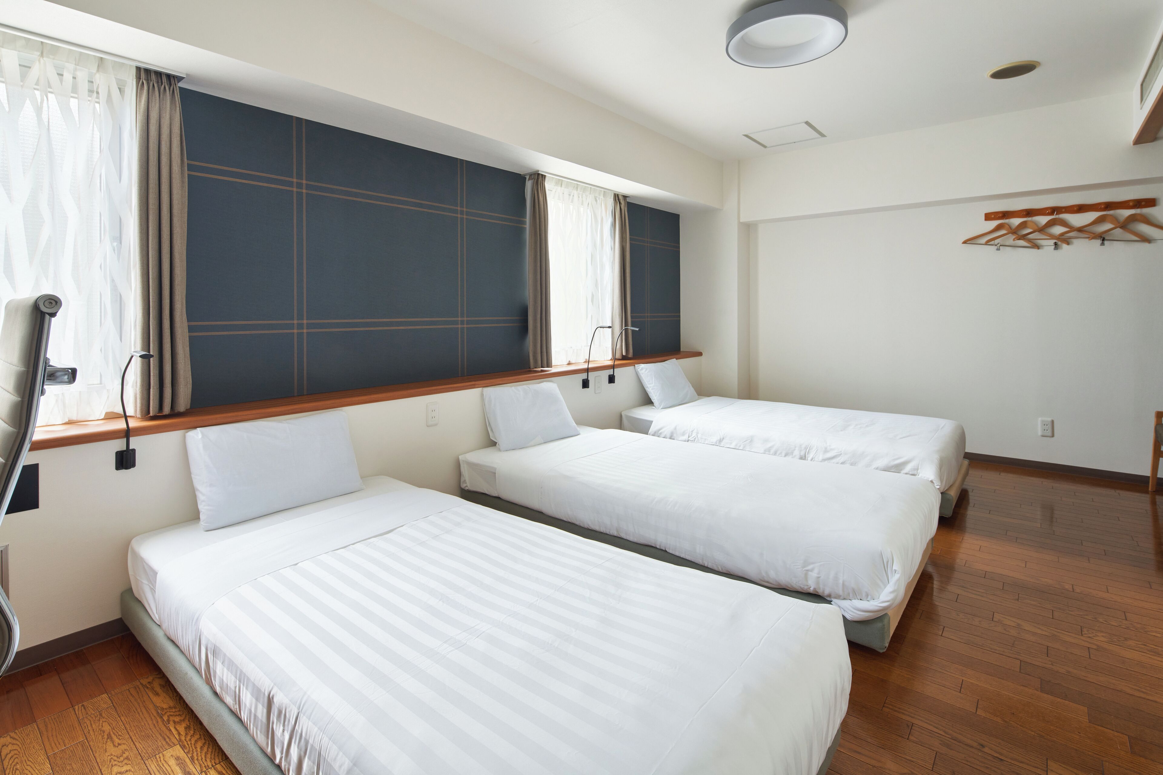 A hotel room with two single beds, a wooden floor, a large window with curtains, and a coat rack.