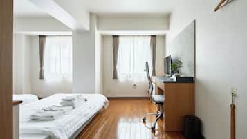 Et hotelværelse med trægulv, seng med hvide sengetøj, skrivebord med computer og et vindue med gardiner.