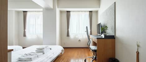 Una habitación de hotel con piso de madera, cama con ropa blanca, escritorio con computadora y ventana con cortinas.