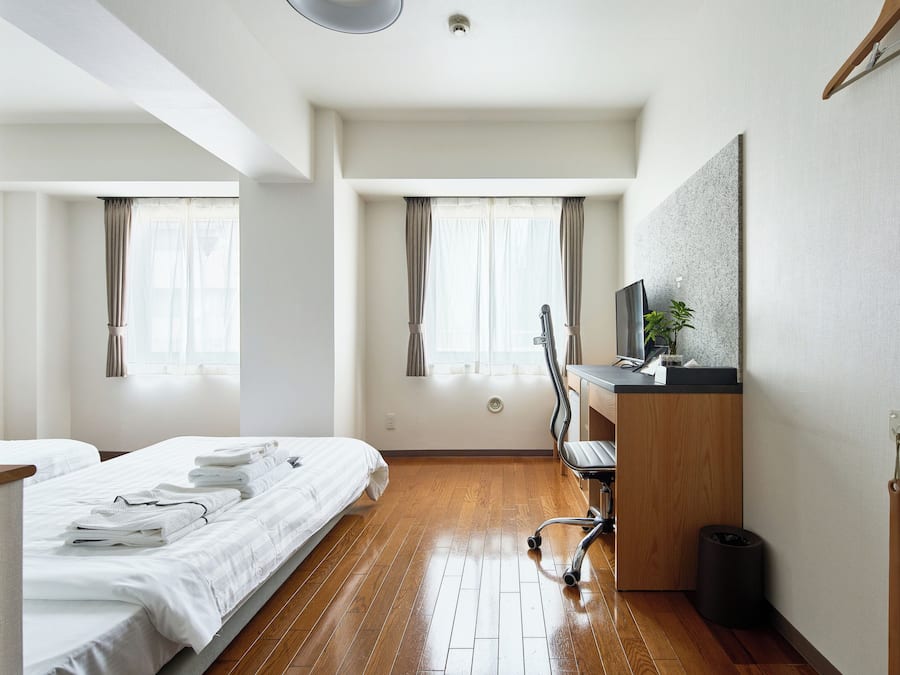 A hotel room with a wooden floor, a bed with white linens, a desk with a computer, and a window with curtains.