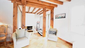 Een woonkamer met een open haard, houten balken en twee witte fauteuils.