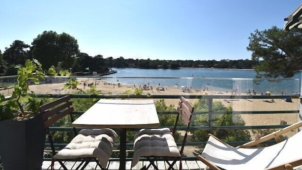 A balcony with a table and two chairs overlooking a beach and lake. - Hossegor “Côte d’Argent”, Apartment on the edge of the lake for 2 people. (HOSSEGOR)