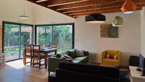 Een ruime woonkamer met een houten plafond, een eettafel met stoelen, een bank, een gele fauteuil en een televisie op een standaard.