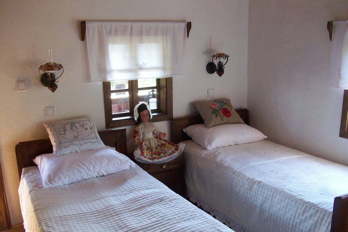 A room with two single beds, a wooden headboard, a window with a white valance, and a wall-mounted lamp.