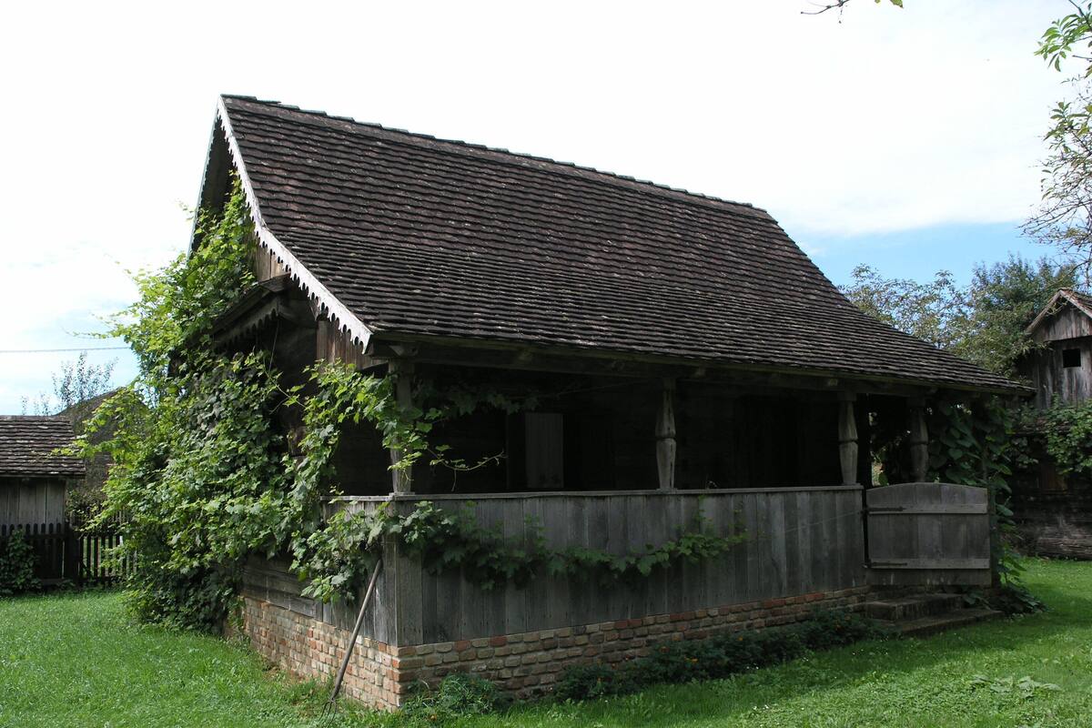 A rustic wooden building with a sloped roof, surrounded by greenery and a brick wall.