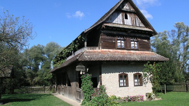 A two-story wooden house with a steeply pitched roof, a balcony, and a stone foundation. - Ethno Village Stara Lonja (Sisak)