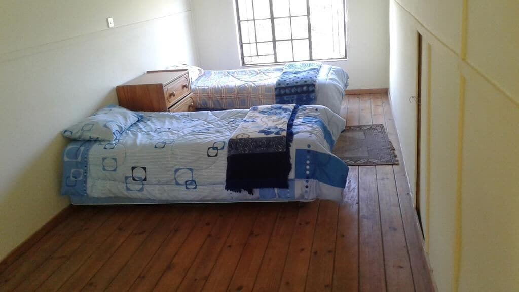 Una habitación pequeña con suelo de madera, dos camas con ropa de cama estampada, un aparador de madera y una ventana con barrotes.