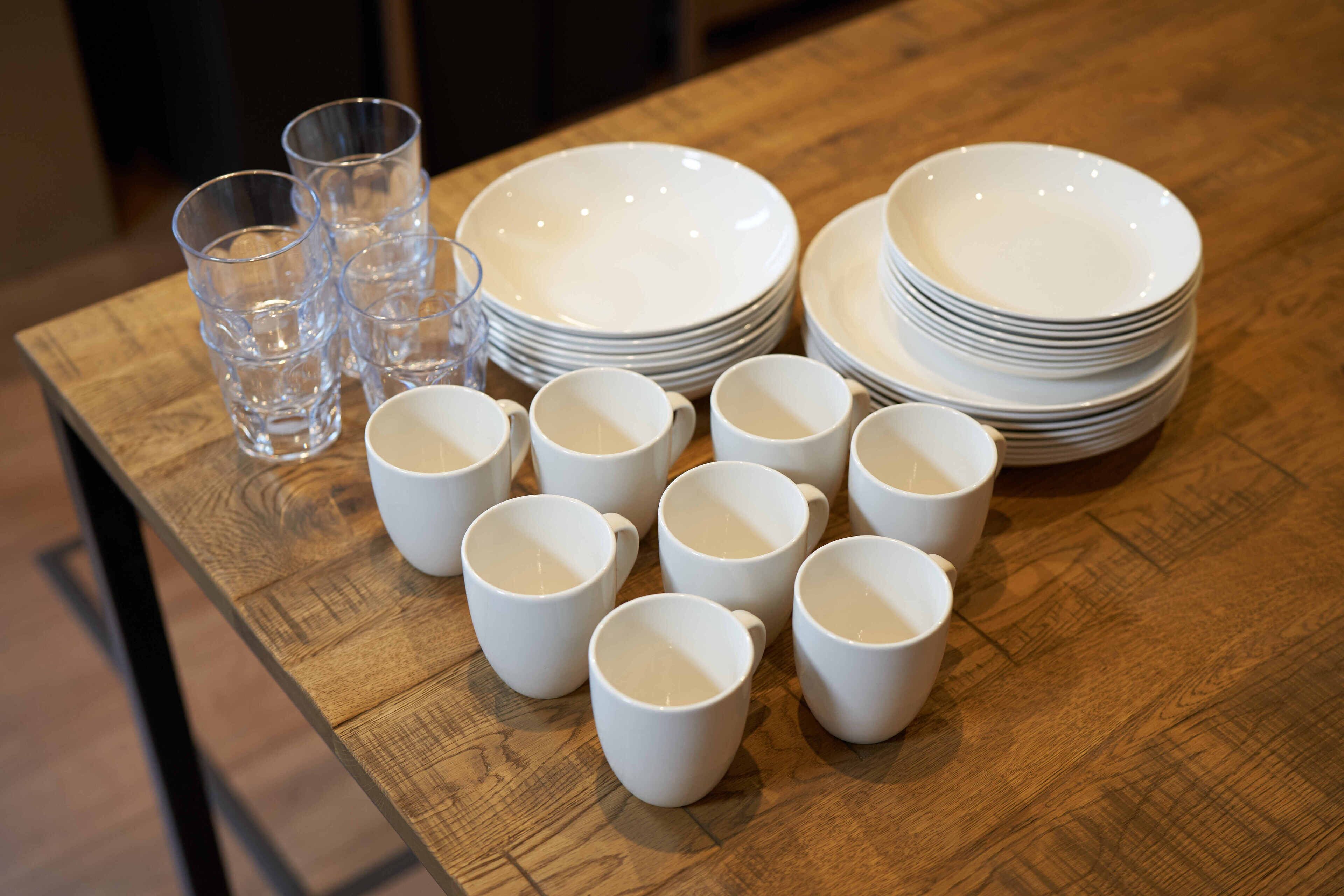 A wooden table with stacks of white plates and cups, and a few clear glasses.