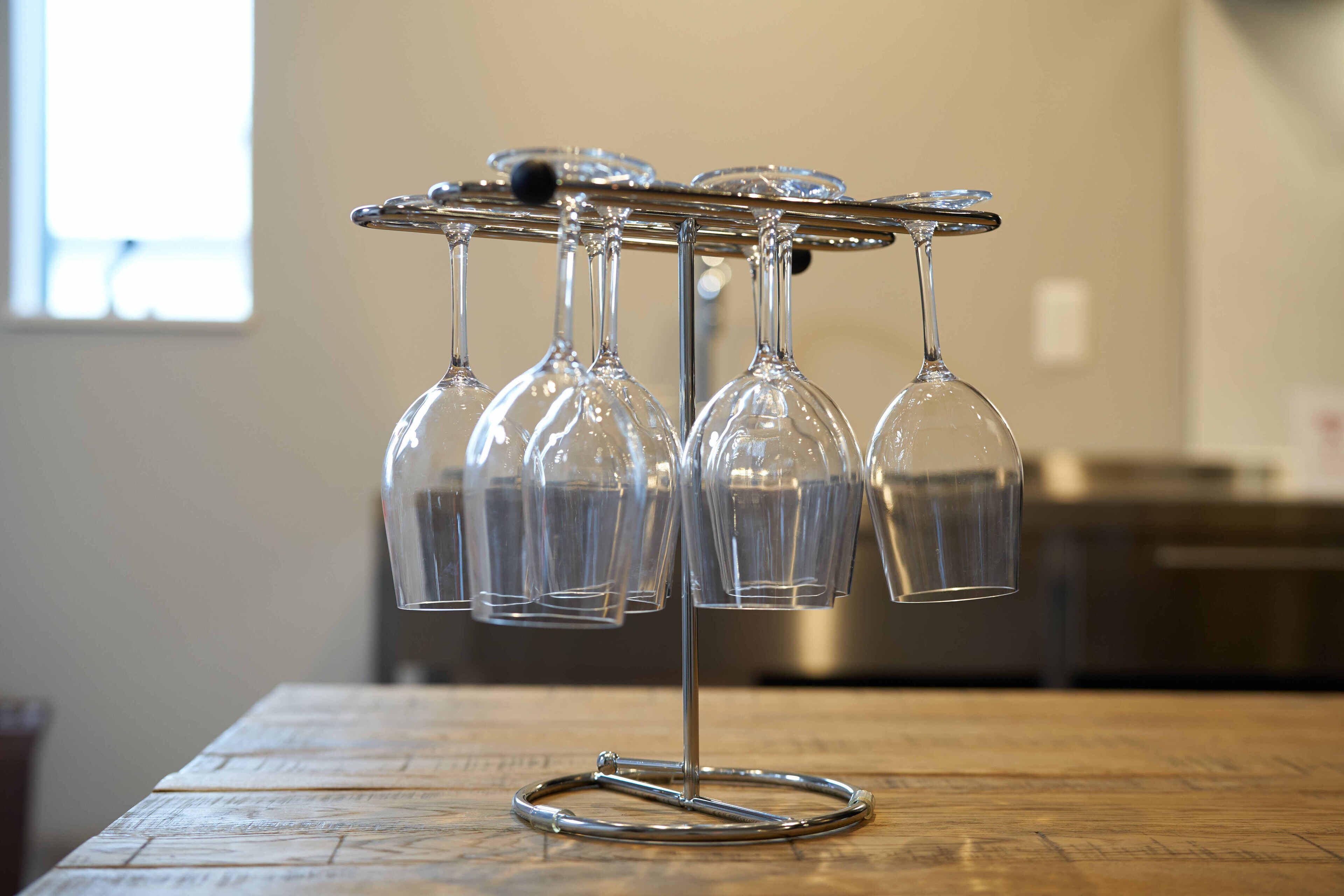 A wine glass holder with six glasses on a metal stand, placed on a wooden table in a room with a window and kitchen appliances in the background.