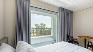 A hotel room with a large bed, a small table, a chair, and a balcony with a view of palm trees and a building. - Swimeong Nolmeong Guesthouse&Pension (Jeju City)