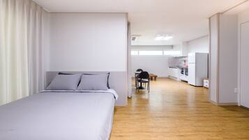A modern hotel room with a large bed, a wooden floor, a kitchenette, and a bathroom visible in the background.