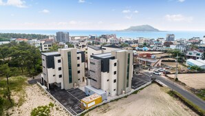 A modern apartment complex with multiple buildings, a parking area, and a view of the sea and a hill in the background. - Swimeong Nolmeong Guesthouse&Pension (Jeju City)