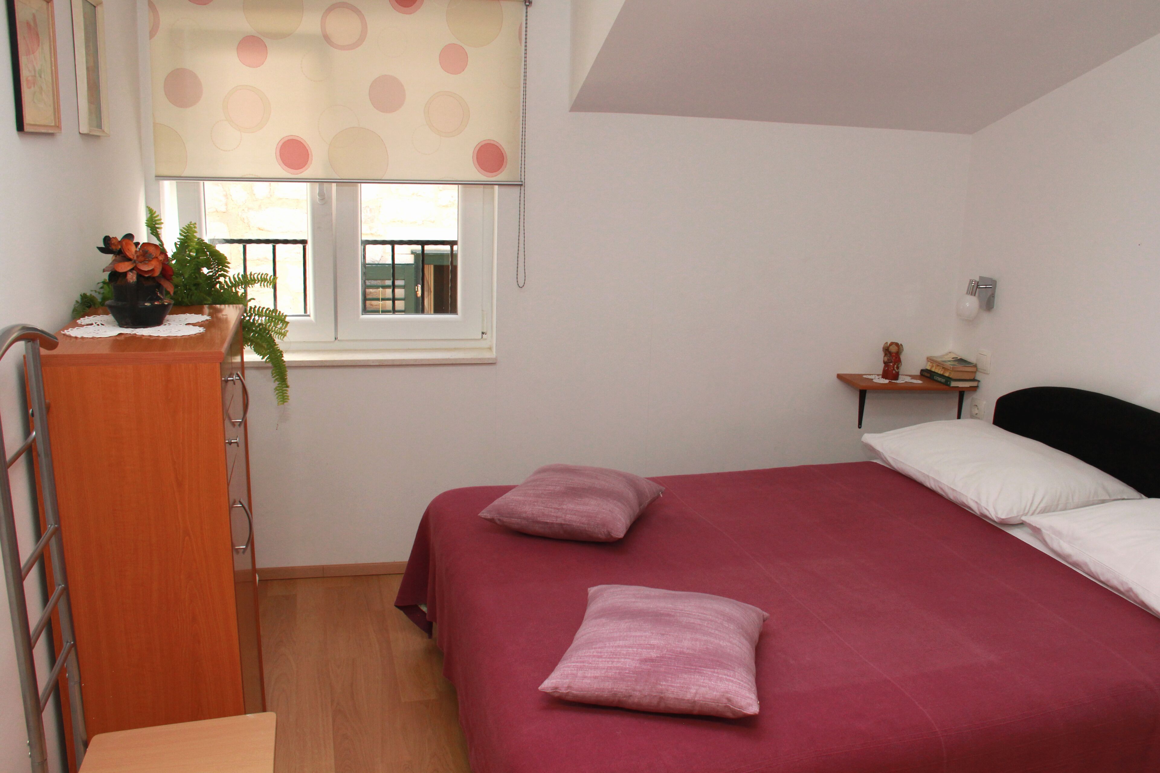 A bedroom with a bed, pillows, a nightstand, a window with a curtain, a desk with a plant, and a small shelf with books.
