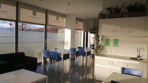 A modern dining area with tables and chairs, a kitchen area with cabinets and a sink, and large windows with frosted glass panels. - Residencia La Ribera (Logroño)
