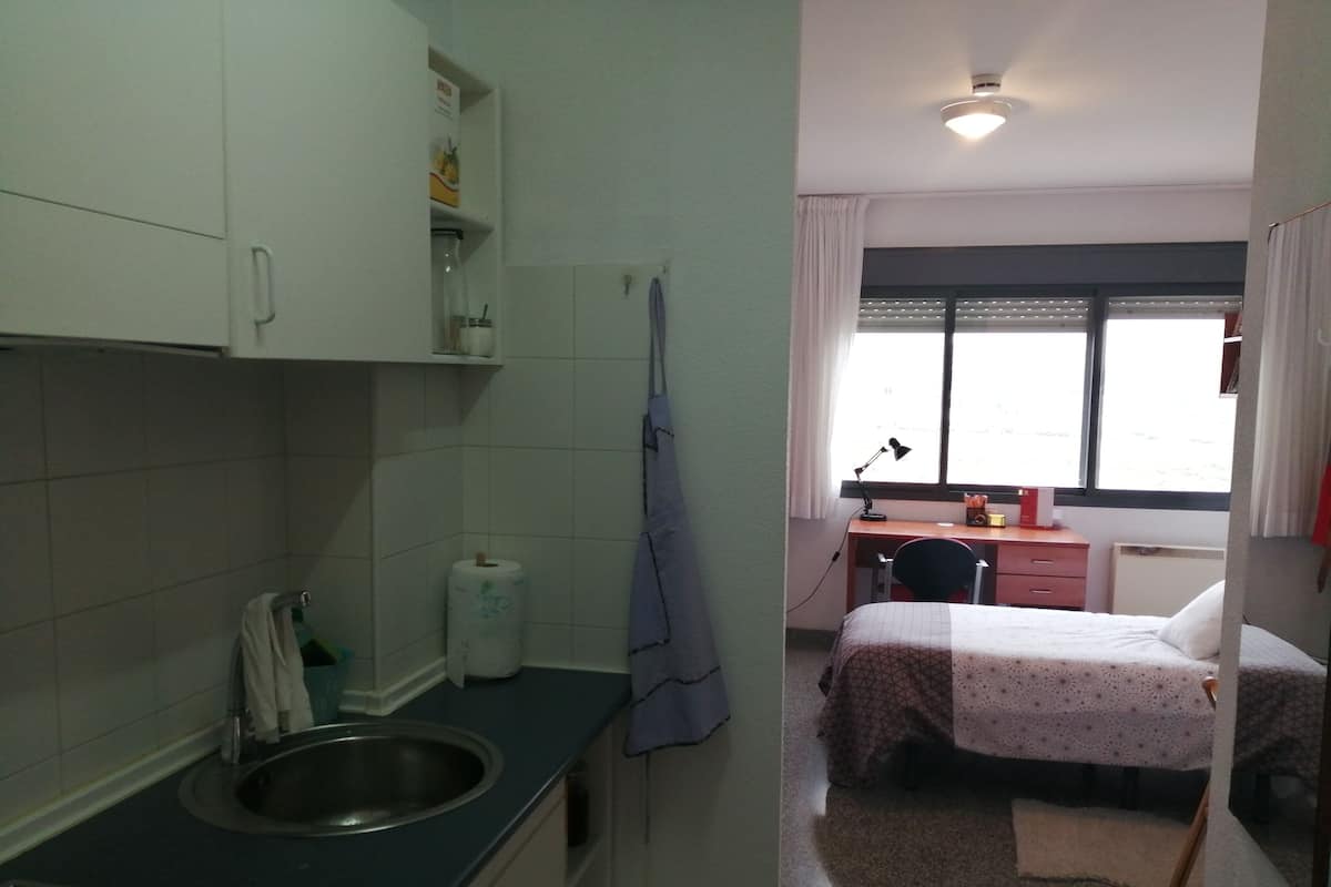 Ein kleines Zimmer mit einer Küchenzeile, einem Bett, einem Schreibtisch und einem Fenster.