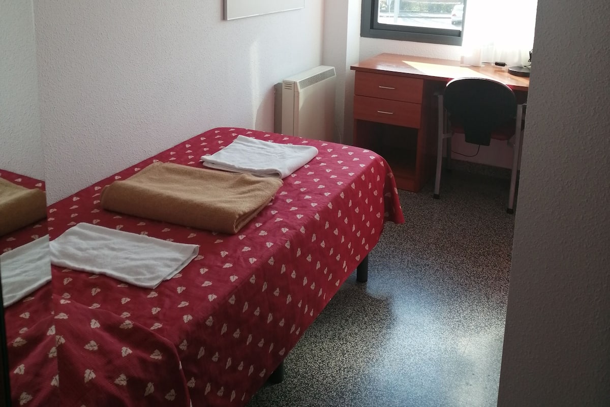 Ein kleiner, ordentlicher Raum mit einem Bett, einem Schreibtisch und einem Fenster.