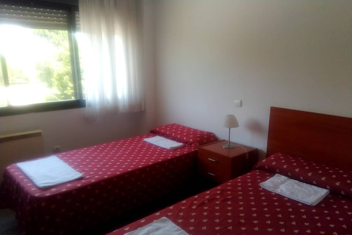 Ein Zimmer mit zwei Einzelbetten, beide mit roter Bettdecke und weißen Kissen. Dazwischen steht ein Nachttisch mit einer Lampe.