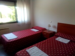 A room with two single beds, each with a red bedspread and white pillows. There is a nightstand with a lamp between the beds. - Residencia La Ribera (Logroño)